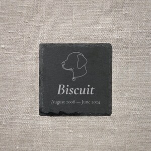 Pode incluir: Uma base de ard&oacute;sia preta quadrada com um contorno branco da cabe&ccedil;a de um c&atilde;o e o nome "Biscuit". Abaixo, as datas "Agosto 2008 &ndash; Junho 2024". A base est&aacute; sobre um fundo de linho neutro.