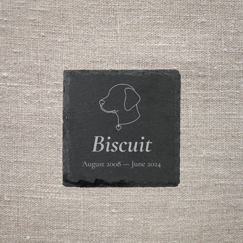 Pode incluir: Uma base de ard&oacute;sia preta quadrada com um contorno branco da cabe&ccedil;a de um c&atilde;o e o nome "Biscuit". Abaixo, as datas "Agosto 2008 &ndash; Junho 2024". A base est&aacute; sobre um fundo de linho neutro.