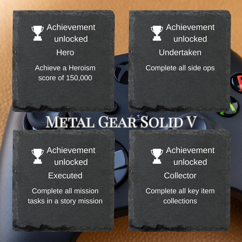 Puede incluir: Cuatro posavasos cuadrados de pizarra con texto blanco e iconos de trofeos. Cada posavasos muestra un logro diferente del videojuego Metal Gear Solid V. Un mando de juego es visible en el fondo.