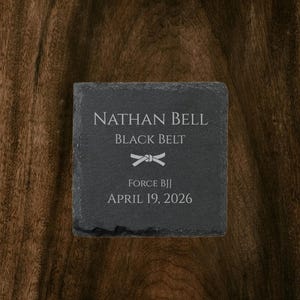 Op de afbeelding: Een vierkante, donkergrijze leistenen onderzetter met de tekst "NATHAN BELL BLACK BELT" en "FORCE BJJ APRIL 19, 2026" in het wit. Een witte afbeelding van een geknoopte riem staat tussen de naam en de BJJ-informatie. De onderzetter ligt op een donkere houten ondergrond.