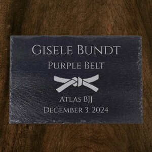 Op de afbeelding: Zwarte leistenen plaquette met witte tekst. De plaquette leest "GISELE BUNDT, PAARSE BAND, ATLAS BJJ, 3 DECEMBER 2024" en heeft een witte bandafbeelding. De achtergrond is een donkere houtnerf.