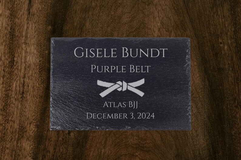 Op de afbeelding: Zwarte leistenen plaquette met witte tekst. De plaquette leest "GISELE BUNDT, PAARSE BAND, ATLAS BJJ, 3 DECEMBER 2024" en heeft een witte bandafbeelding. De achtergrond is een donkere houtnerf.
