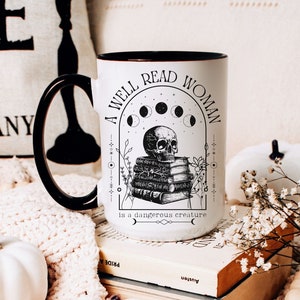 K&ouml;nnte beinhalten: Eine wei&szlig;e Keramiktasse mit schwarzem Rand und Henkel. Die Tasse zeigt eine Schwarzwei&szlig;illustration eines Sch&auml;dels, der auf einem Stapel B&uuml;cher sitzt, mit einer zunehmenden Mondsichel und Sternen dar&uuml;ber. Der Text lautet "Eine Frau, die viel liest, ist eine gef&auml;hrliche Kreatur."