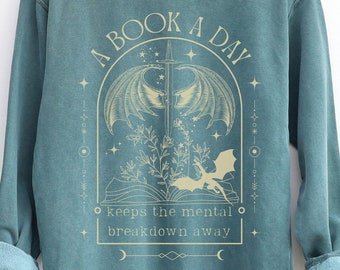 May include: A teal blue sweatshirt with a graphic design of a book with wings and a sword. The text reads "A Book A Day keeps the mental breakdown away".