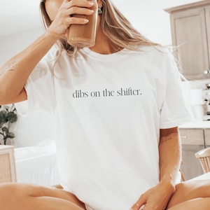 May include: A white t-shirt with the text "dibs on the shifter." printed on the front. The shirt is being worn by a person sitting on a white surface.