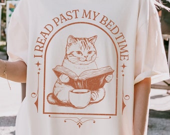 May include: A white t-shirt with a brown graphic of a cat reading a book and drinking coffee. The text on the shirt reads "I read past my bedtime".