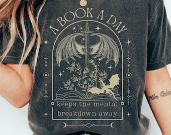 May include: A dark gray t-shirt with a graphic design of a book with a sword and two dragons. The text "A Book A Day keeps the mental breakdown away" is printed on the shirt.