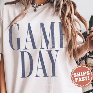 May include: An ivory short-sleeved t-shirt featuring the text GAME DAY in large, distressed navy blue capital letters. The shirt has a crew neckline and a relaxed fit. The word IVORY is printed in small letters at the bottom centre.