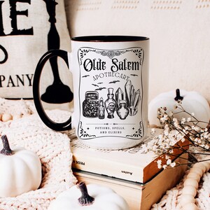 May include: White ceramic mug with a black handle and black rim. The mug has a black and white graphic of a vintage apothecary label with the text "Olde Salem Apothecary" and the words "Potions, Spells, and Elixirs". The graphic also includes illustrations of bottles, crystals, and bats.