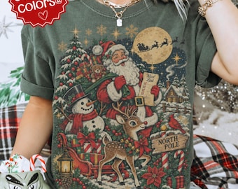 Könnte beinhalten: Ein dunkelolivgrünes T-Shirt mit einer detaillierten Weihnachtsgrafik, die den Weihnachtsmann mit einer Liste, einen Schneemann, ein Rentier, einen Schlitten mit Geschenken, ein Nordpol-Schild und einen Vollmond mit Schlittensilhouette zeigt.