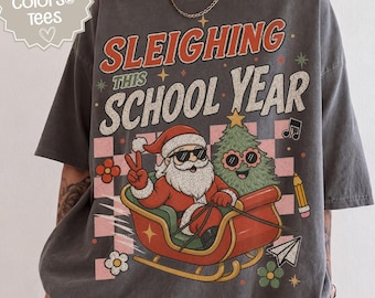 May include: A charcoal grey t-shirt featuring a graphic of Santa Claus in sunglasses riding a red sleigh with a decorated Christmas tree. The text reads "SLEIGHING THIS SCHOOL YEAR" with icons of flowers, a pencil, and a paper airplane on a checkered background.