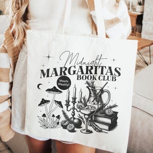 May include: White canvas tote bag with black text and illustrations. The text reads "Midnight Margaritas Book Club" with the words "Meets Weekly!" in a smaller font. The illustration features a pitcher of margaritas, a glass of margarita, mushrooms, and a candelabra.