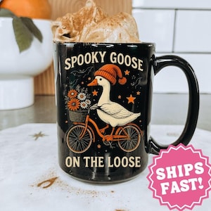 May include: Black 15 oz ceramic mug with a whimsical design. The mug features a goose wearing a red hat and riding a bicycle, with the words "Spooky Goose on the Loose". The mug has a black handle.