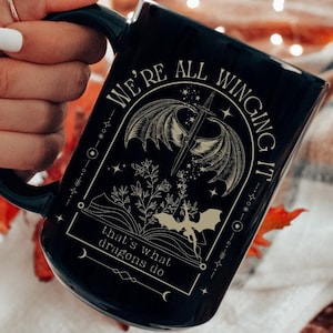 May include: Black ceramic mug with a gold design featuring a dragon with wings spread, a sword, and a book. The text reads "We're all winging it that's what dragons do."