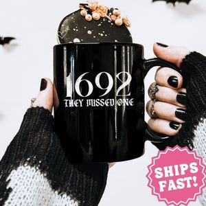 May include: Black ceramic mug with the text "1692 THEY MISSED ONE" in white. The mug is held by a person wearing a black and white sweater. A decorative cookie sits on top of the mug. The mug is 11 oz.