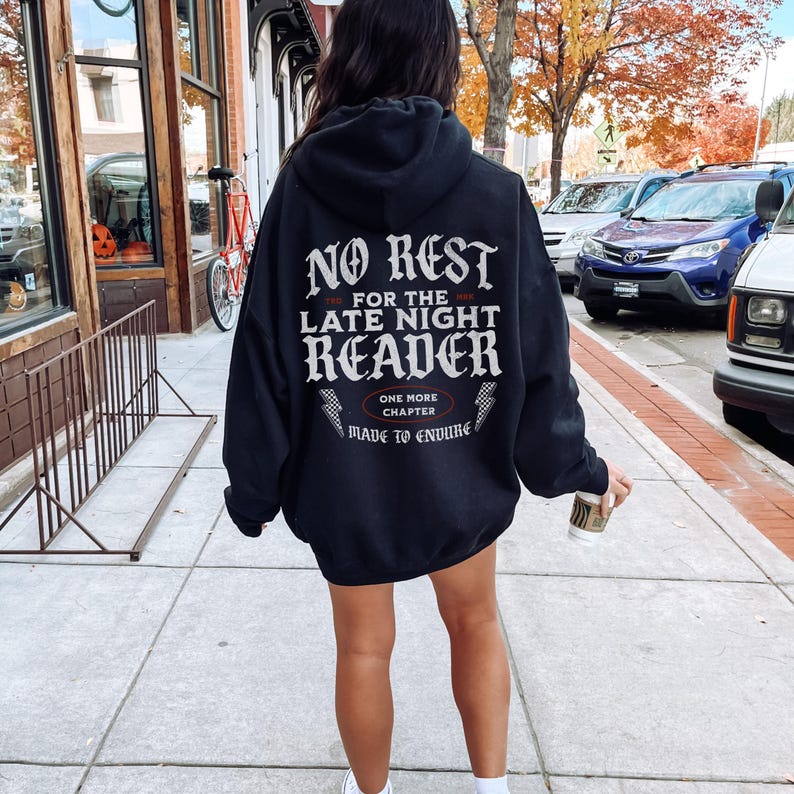 Peut inclure: Un sweat &agrave; capuche noir avec un texte blanc au dos : "NO REST for the LATE NIGHT READER", avec "ONE MORE CHAPTER" et "MADE TO ENDURE" en dessous. La personne porte le sweat &agrave; capuche et marche sur un trottoir.