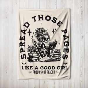 May include: A beige throw blanket with a black and white illustration of a skeleton reading a book. The text on the blanket reads "Spread those pages like a good girl, Proud Smut Reader."