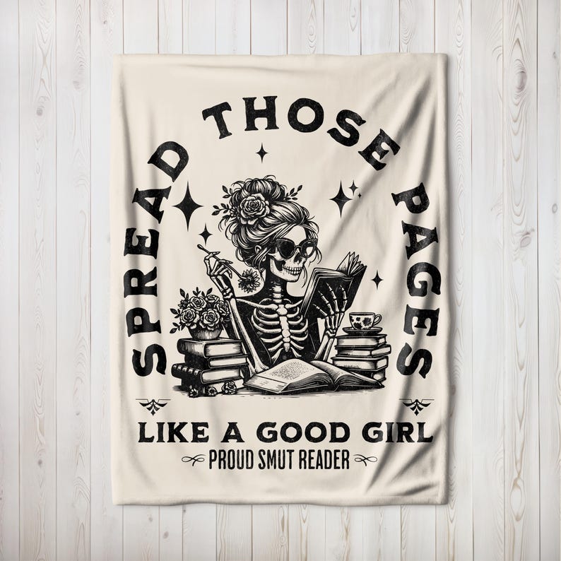 May include: A beige throw blanket with a black and white illustration of a skeleton reading a book. The text on the blanket reads "Spread those pages like a good girl, Proud Smut Reader."