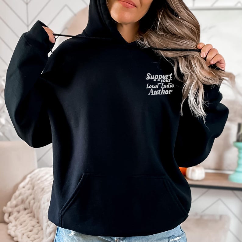 May include: Black hooded sweatshirt with a front pocket and drawstring. The text "Support Your Local Indie Author" is printed in white script on the upper chest. The model is wearing blue jeans.