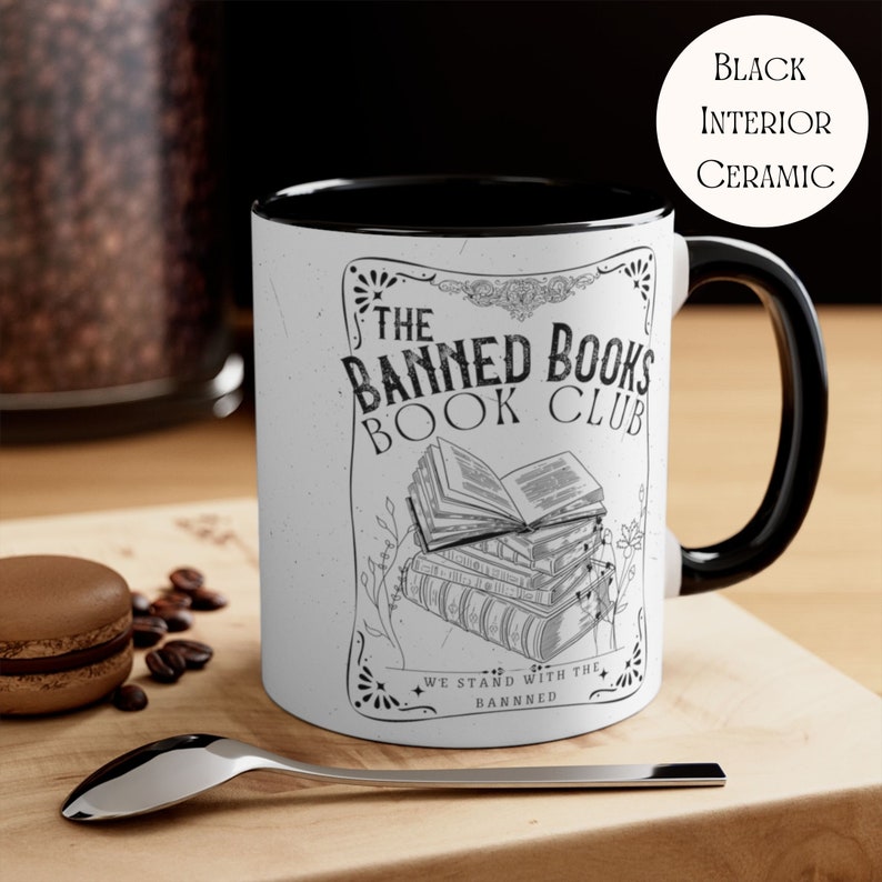 Op de afbeelding: Een witte keramische mok met een zwarte binnenkant en handvat. De mok heeft een zwart-witte illustratie van een stapel boeken met de tekst "The Banned Books Book Club" en "We Stand With The Banned".