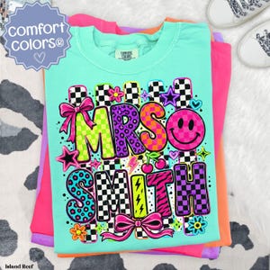 May include: A mint green t-shirt featuring the text MRS SMITH in colorful, patterned block letters. The design includes black and white checkerboard accents, pink bows, a smiley face, stars, lightning bolts, and cherries on a bright background.