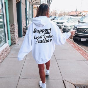 May include: A white hoodie with the text "Support your Local Indie Author" printed in black on the back. The hoodie is oversized and worn by a person walking on a sidewalk.
