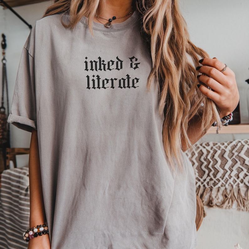 May include: A gray t-shirt with the words "inked & literate" in a gothic font. The shirt has short sleeves and a relaxed fit. The text is in black.