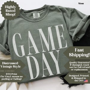 May include: A moss green short-sleeve t-shirt featuring the text GAME DAY printed in large, white, distressed-style capital letters across the chest. The shirt has a crew neckline and a visible brand label on the inside of the collar.