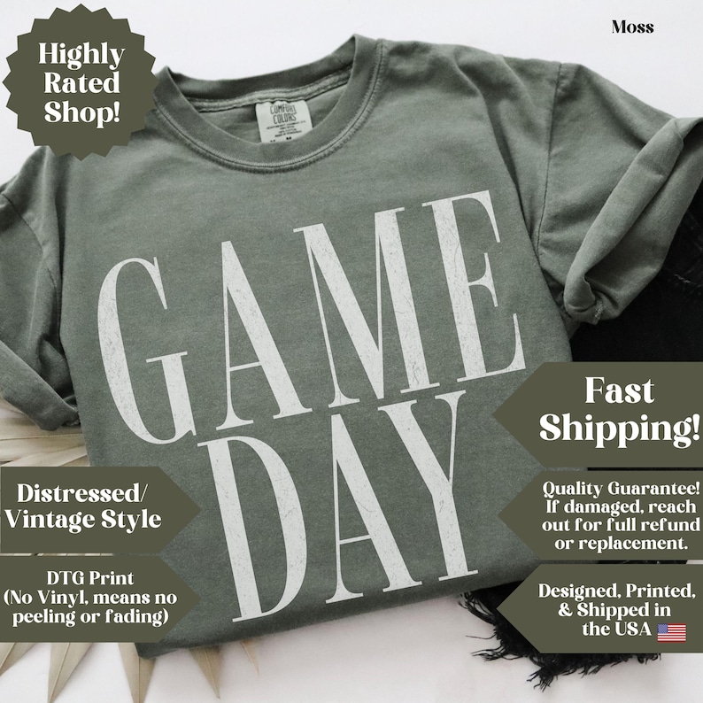 May include: A moss green short-sleeve t-shirt featuring the text GAME DAY printed in large, white, distressed-style capital letters across the chest. The shirt has a crew neckline and a visible brand label on the inside of the collar.