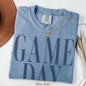May include: A blue jean coloured short-sleeve t-shirt with a crew neckline. The shirt features the text GAME DAY printed in large, dark blue, distressed capital letters across the front. A Comfort Colors brand label is visible at the inner neckline.