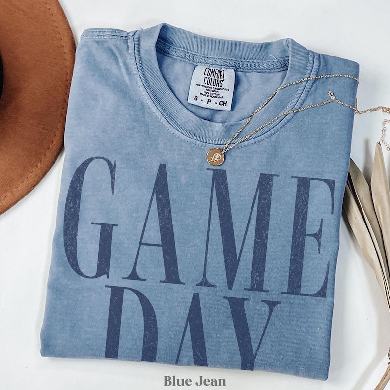 May include: A blue jean coloured short-sleeve t-shirt with a crew neckline. The shirt features the text GAME DAY printed in large, dark blue, distressed capital letters across the front. A Comfort Colors brand label is visible at the inner neckline.