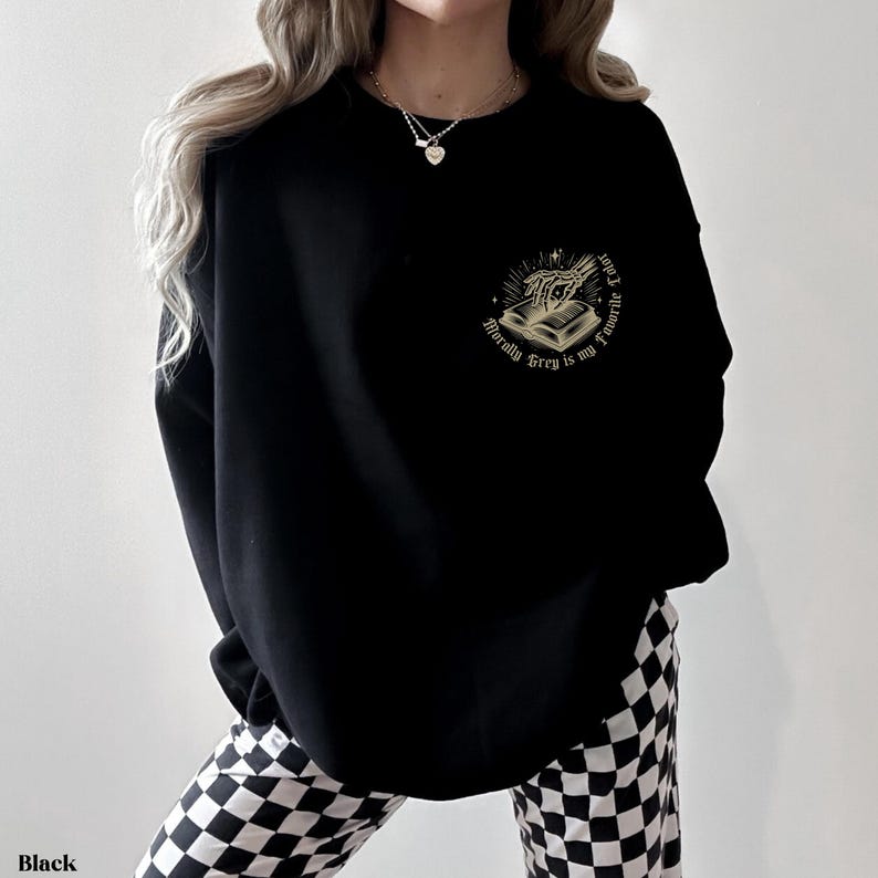 May include: Black sweatshirt with a gold graphic on the upper left chest. The graphic features a skeleton hand holding a book with the text "Morally Grey is my Favorite". The sweatshirt is a solid black color.