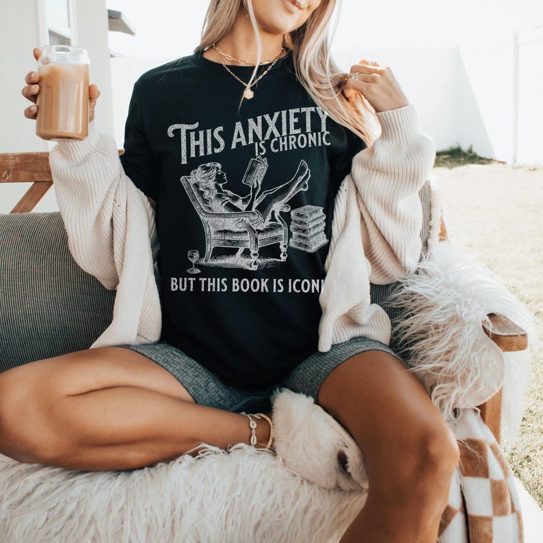 K&ouml;nnte beinhalten: Schwarzes T-Shirt mit einer wei&szlig;en Grafik einer Frau, die in einem Sessel ein Buch liest. Der Text auf dem Shirt lautet "This anxiety is chronic but this book is iconic."