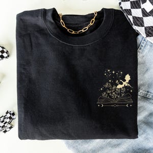 May include: Black sweatshirt with a gold graphic of a dragon flying out of an open book with stars and crescent moons.