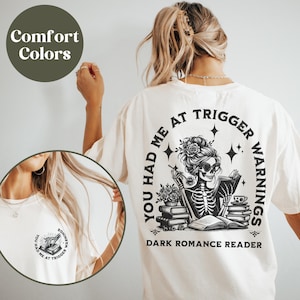 May include: White t-shirt with a black graphic of a skeleton reading a book. The graphic includes the text "You had me at trigger warnings" and "Dark Romance Reader".