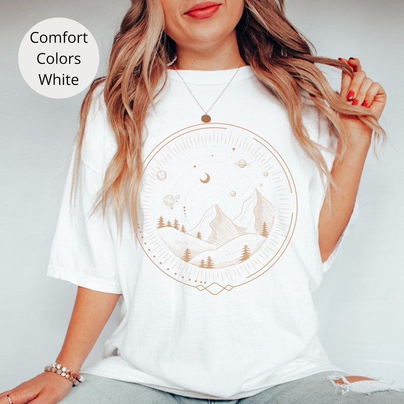 Puede incluir: Camiseta blanca con un dibujo de l&iacute;nea marr&oacute;n de monta&ntilde;as, una luna creciente y estrellas dentro de un c&iacute;rculo. El dise&ntilde;o recuerda un paisaje celestial.