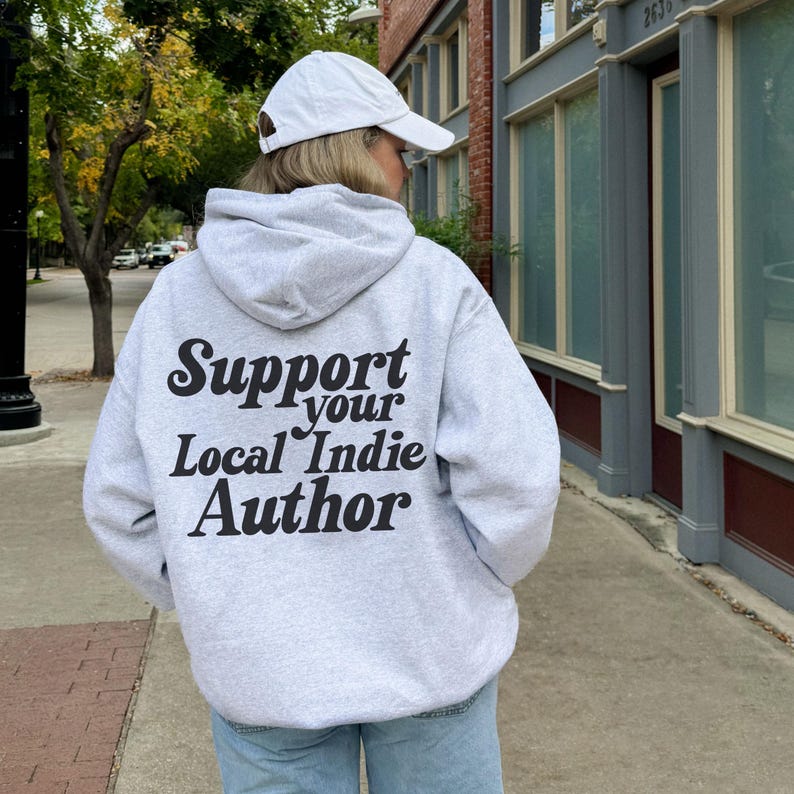 May include: A light gray hoodie with the text "Support your Local Indie Author" in black script. The hoodie has a drawstring hood and is worn with a white baseball cap. The person is standing on a sidewalk.