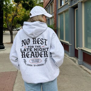 Peut inclure: Un sweat &agrave; capuche gris clair avec le texte "NO REST, LATE NIGHT READER" en noir, en police gothique. Le sweat &agrave; capuche porte &eacute;galement le texte "ONE MORE CHAPTER, MADE TO ENDURE". La personne porte une casquette de baseball blanche et un jean bleu clair.