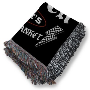 May include: Black woven blanket with white text and a checkered lightning bolt design. The blanket has a fringed edge with multiple colors. The text on the blanket reads "...E'S BLANKET".