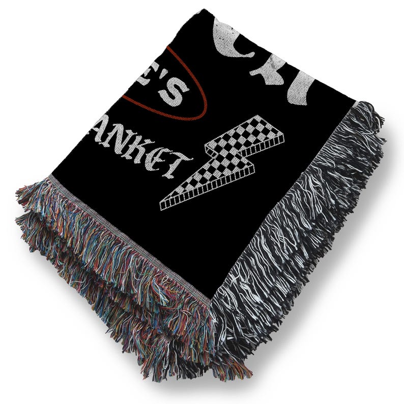 May include: Black woven blanket with white text and a checkered lightning bolt design. The blanket has a fringed edge with multiple colors. The text on the blanket reads "...E'S BLANKET".