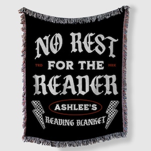 May include: Black throw blanket with white text that reads "NO REST FOR THE READER" and "ASHLEE'S READING BLANKET". The blanket has a fringed edge with red, white, and black threads. Two lightning bolts with a checkered pattern are on the bottom.
