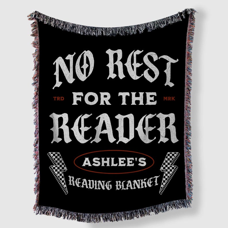 May include: Black throw blanket with white text that reads "NO REST FOR THE READER" and "ASHLEE'S READING BLANKET". The blanket has a fringed edge with red, white, and black threads. Two lightning bolts with a checkered pattern are on the bottom.
