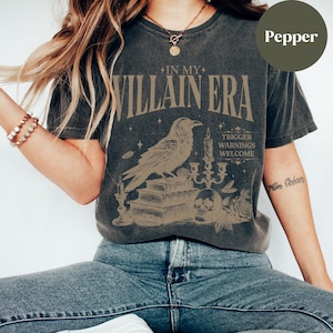 May include: A dark grey t-shirt with a vintage-style graphic featuring a raven perched on a stack of books, a skull, candles, and the text "In My Villain Era. Trigger Warnings Welcome."