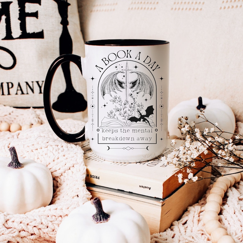 May include: White ceramic mug with a black rim and handle. The mug features a black and white illustration of a book with wings, a sword, and a dragon. The text reads "A Book A Day keeps the mental breakdown away".