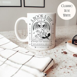 May include: White ceramic mug with a black and white illustration of a book, a sword, and a dragon. The text reads "A Book A Day keeps the mental breakdown away".