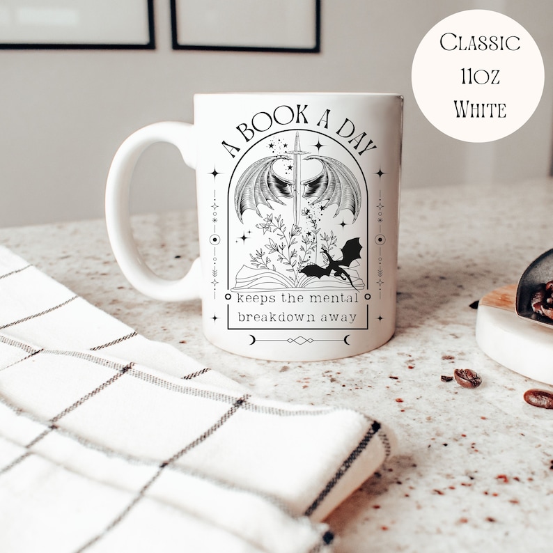 May include: White ceramic mug with a black and white illustration of a book, a sword, and a dragon. The text reads "A Book A Day keeps the mental breakdown away".