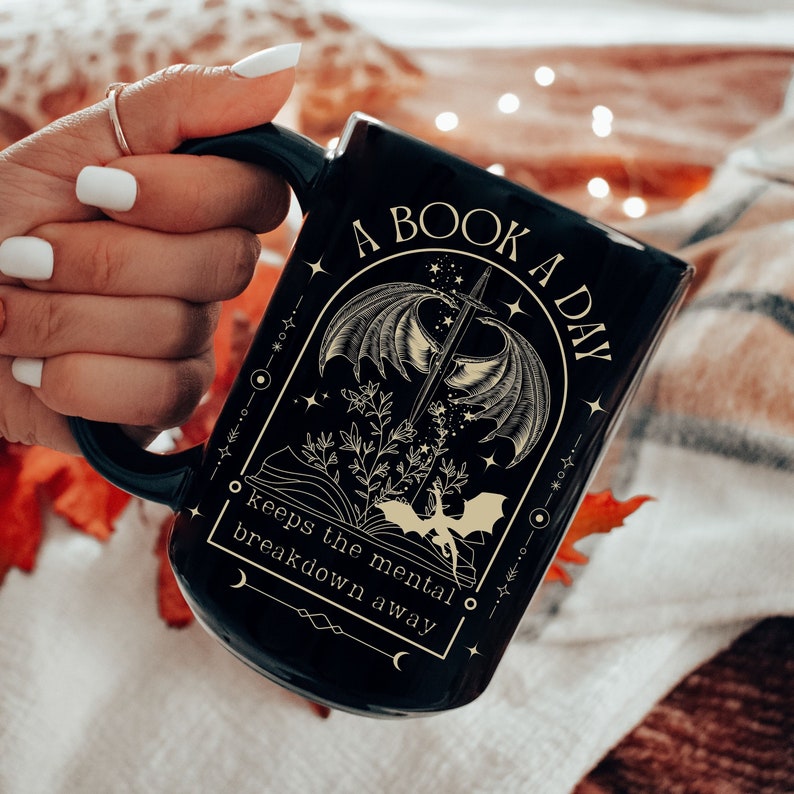 May include: Black coffee mug with a gold design featuring a dragon, a sword, and a book. The text reads "A Book A Day Keeps The Mental Breakdown Away".