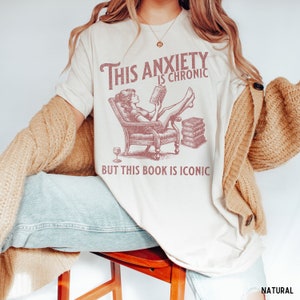 K&ouml;nnte beinhalten: Ein wei&szlig;es T-Shirt mit einer pinkfarbenen Grafik einer Frau, die in einem Stuhl sitzt und ein Buch liest. Der Text auf dem Shirt lautet "This anxiety is chronic but this book is iconic".
