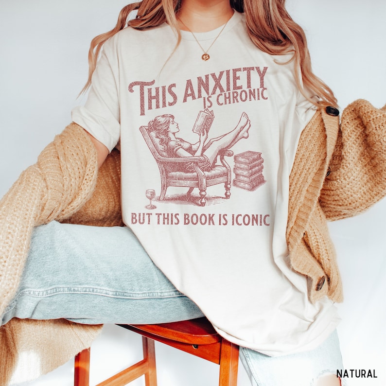 K&ouml;nnte beinhalten: Ein wei&szlig;es T-Shirt mit einer pinkfarbenen Grafik einer Frau, die in einem Stuhl sitzt und ein Buch liest. Der Text auf dem Shirt lautet "This anxiety is chronic but this book is iconic".