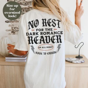 May include: White t-shirt with black text that reads "NO REST FOR THE DARK ROMANCE READER" with a red circle around the text "UP ALL NIGHT" and black and white lightning bolts on either side. The text "MADE TO ENDURE" is below the circle.